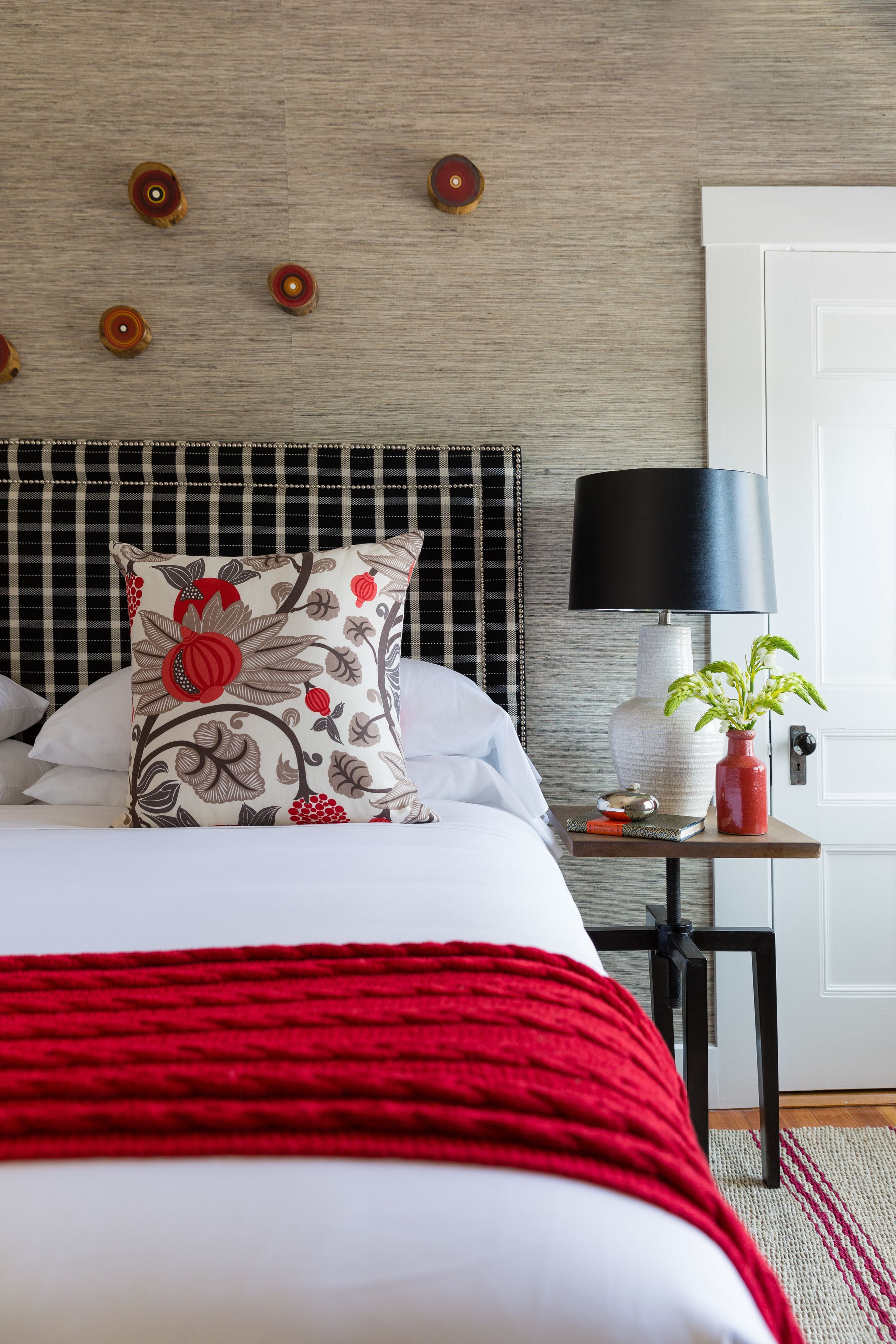 Una cama bien hecha con una colcha roja, una mesita de noche con una lámpara y una planta en maceta, y una pared con luces decorativas redondas.