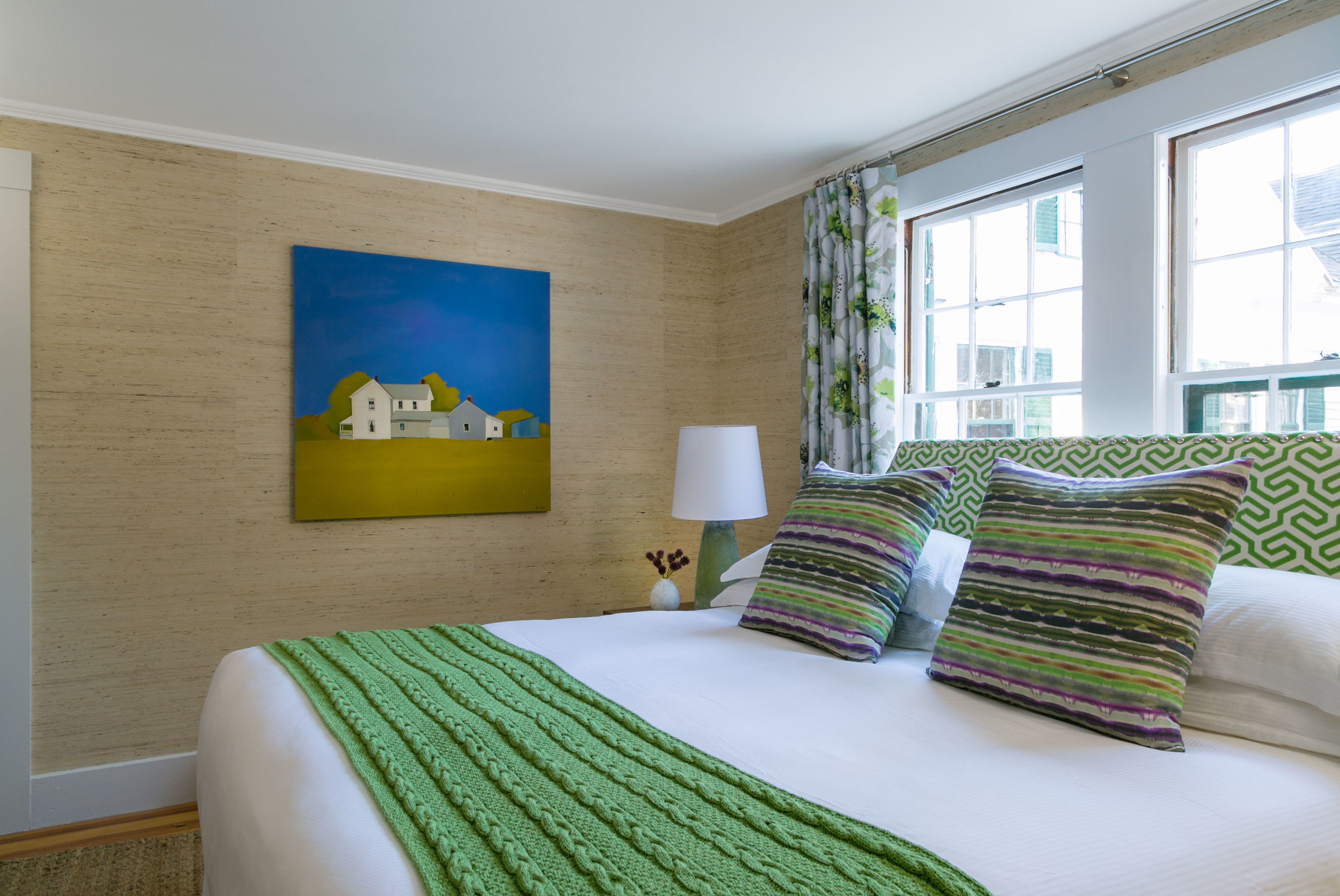 A neatly made bed with a green blanket, a painting of a house on the wall, and a window with curtains.