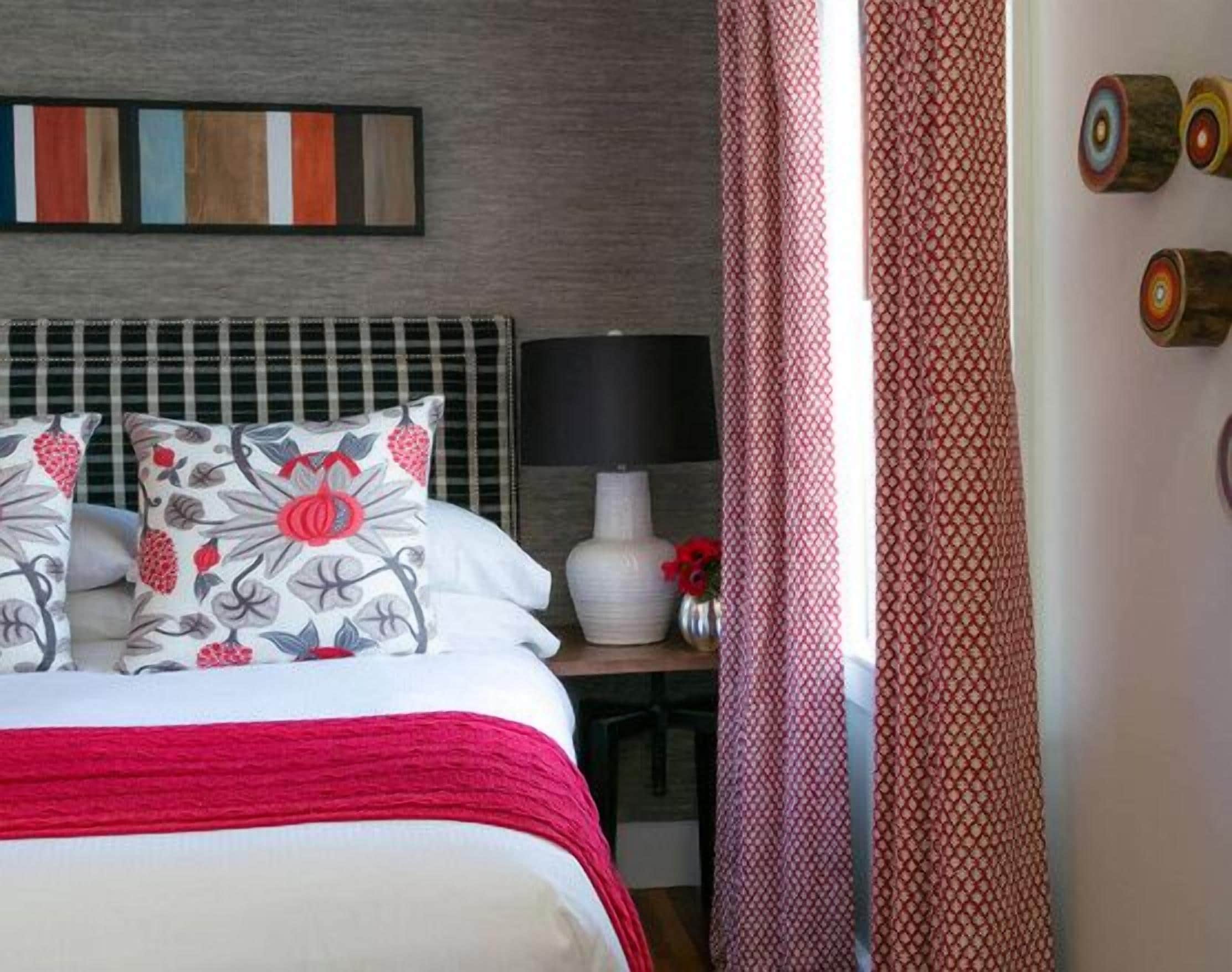 Una cama bien hecha con almohadillas florales, una manta roja y un cabecero moderno. Se ve una mesita de noche con una lámpara y un florero.