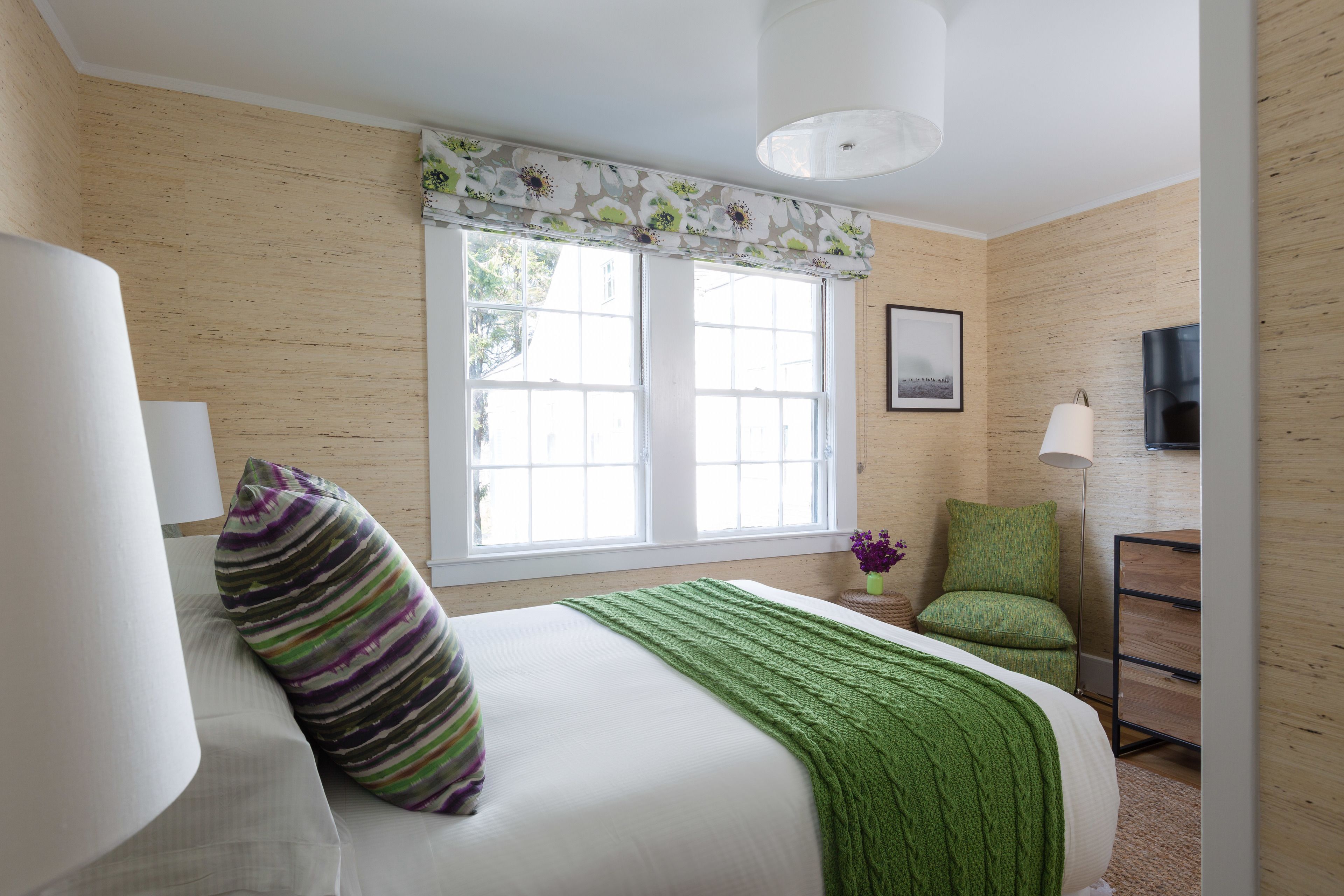 A bedroom with a bed, a green blanket, a striped pillow, a chair, a desk with a lamp, and a window with a view of the outside.