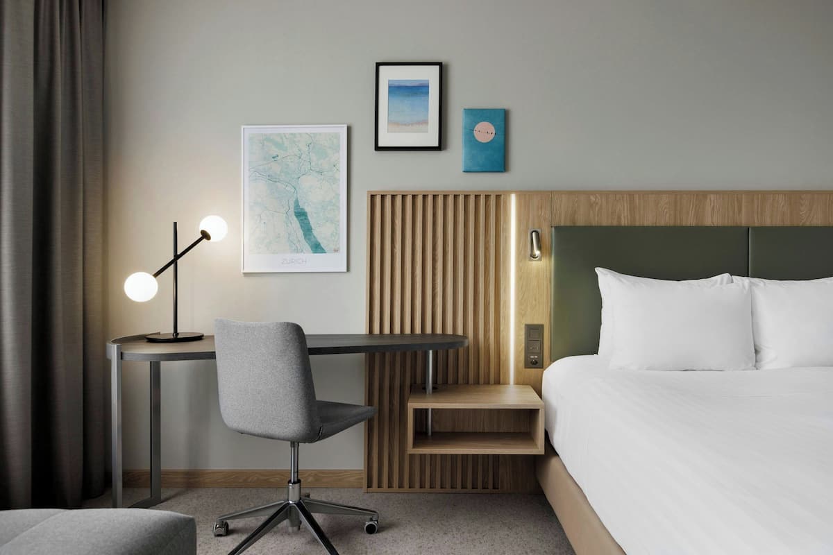 Habitación de hotel con cama, escritorio y silla, lámpara y dos cuadros en la pared.