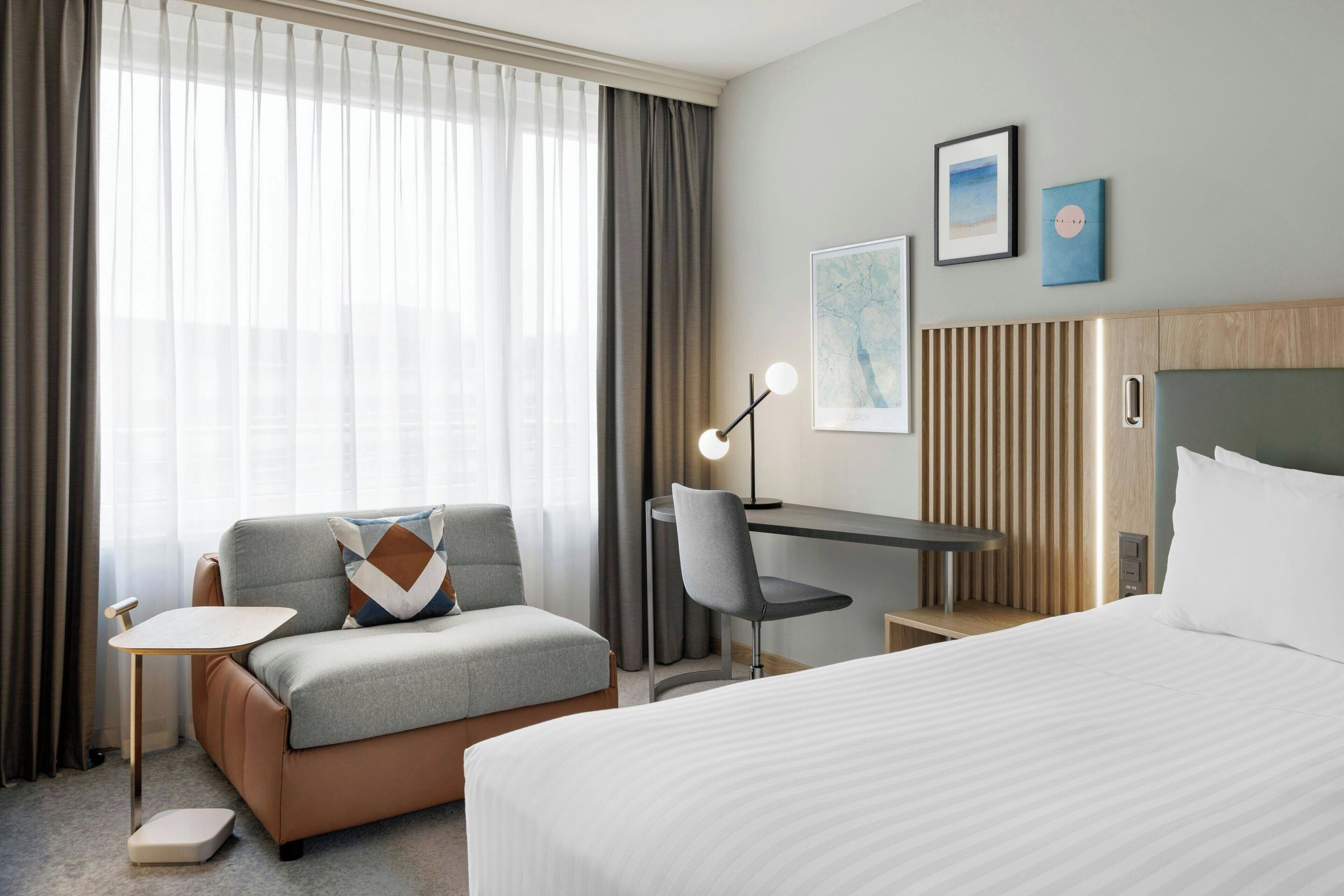 Habitación de hotel con cama, sofá, escritorio y silla. Cuenta con ventana con cortinas, lámpara de pared y dos cuadros enmarcados.