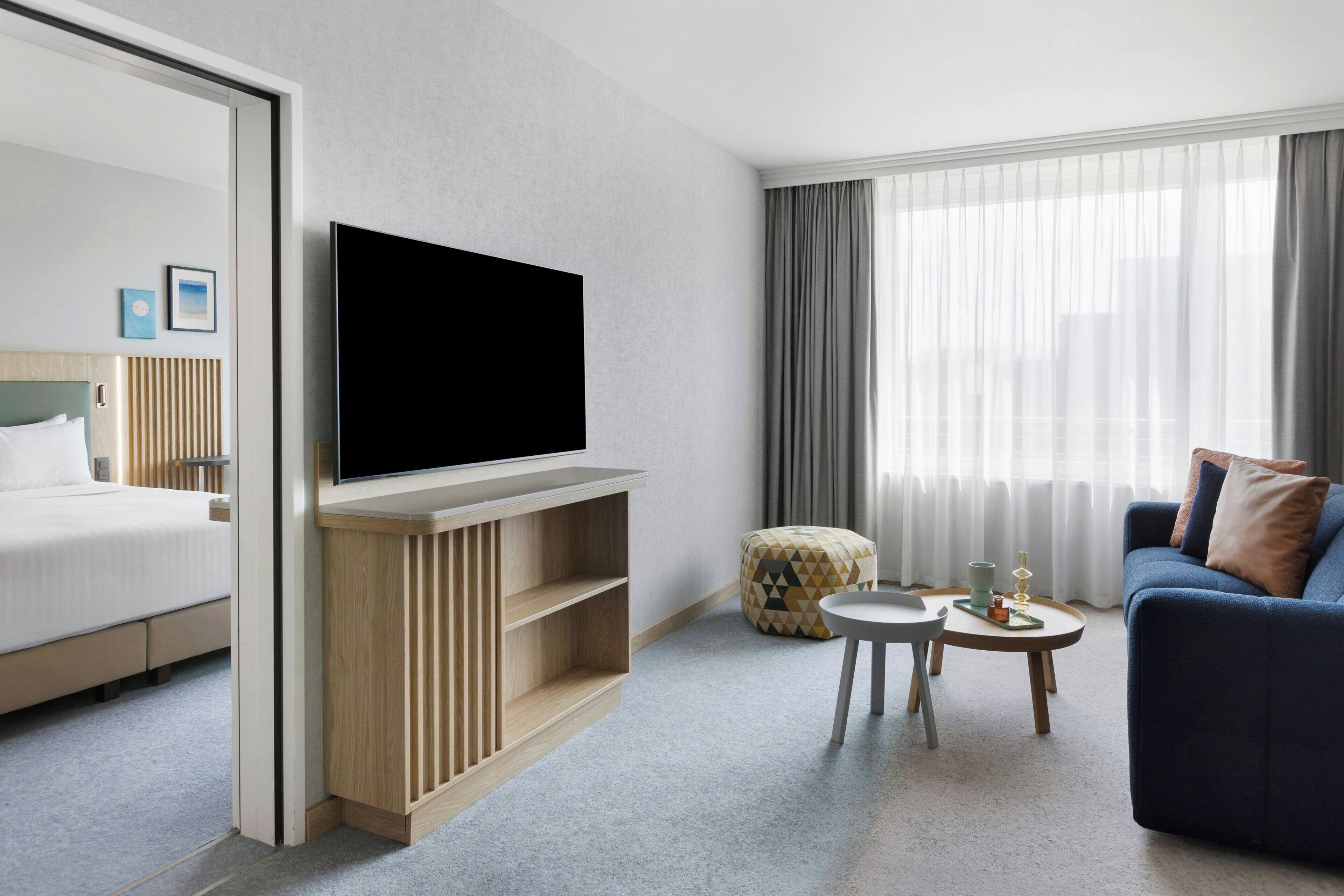 Une chambre d’hôtel moderne équipée d’un lit, d’un téléviseur à écran plat fixé sur une console en bois, d’un canapé, d’une petite table ronde avec un plateau et d’une fenêtre agrémentée de rideaux légers.