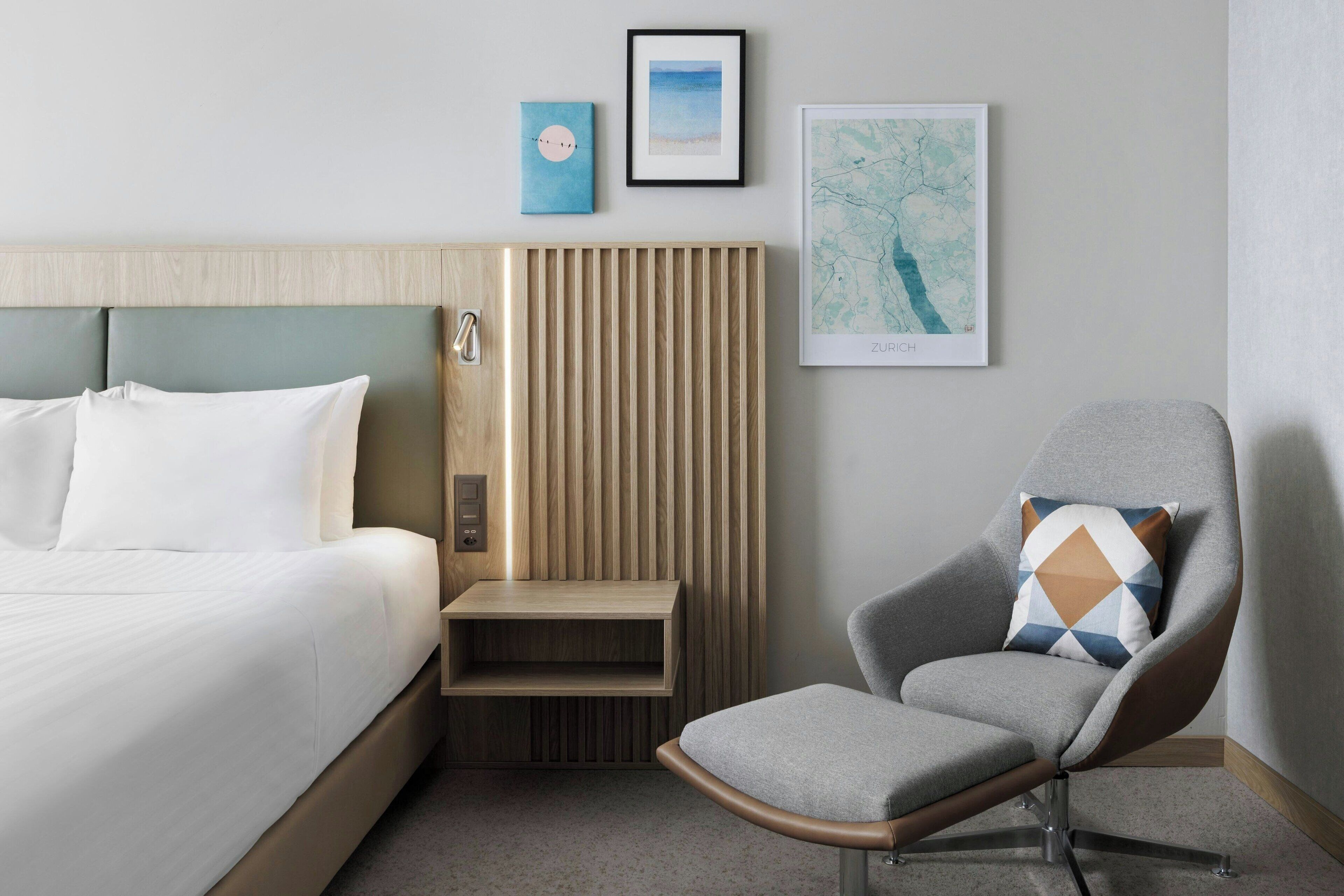 A modern hotel room with a bed, a wooden nightstand, a gray armchair, and wall art.