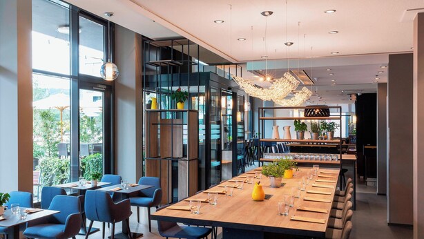 Un restaurante moderno con una mesa común alargada, sillas azules y varias plantas.