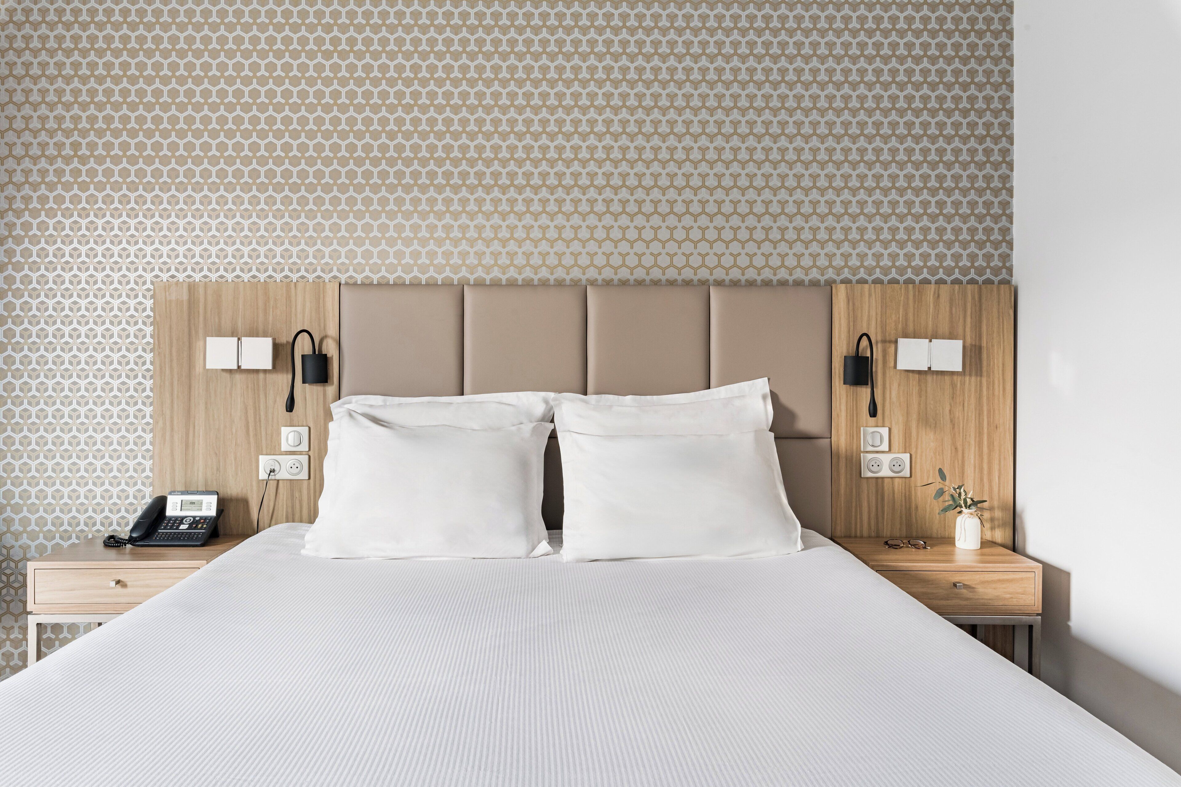 A neatly made bed with two pillows, a bedside table with a phone, a vase with a plant, and a wall with a geometric pattern.