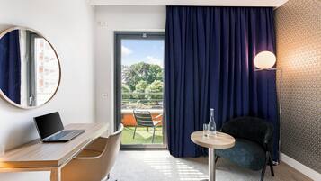 Habitación de hotel con escritorio, laptop, silla, mesa redonda y vista a zonas verdes por la ventana.