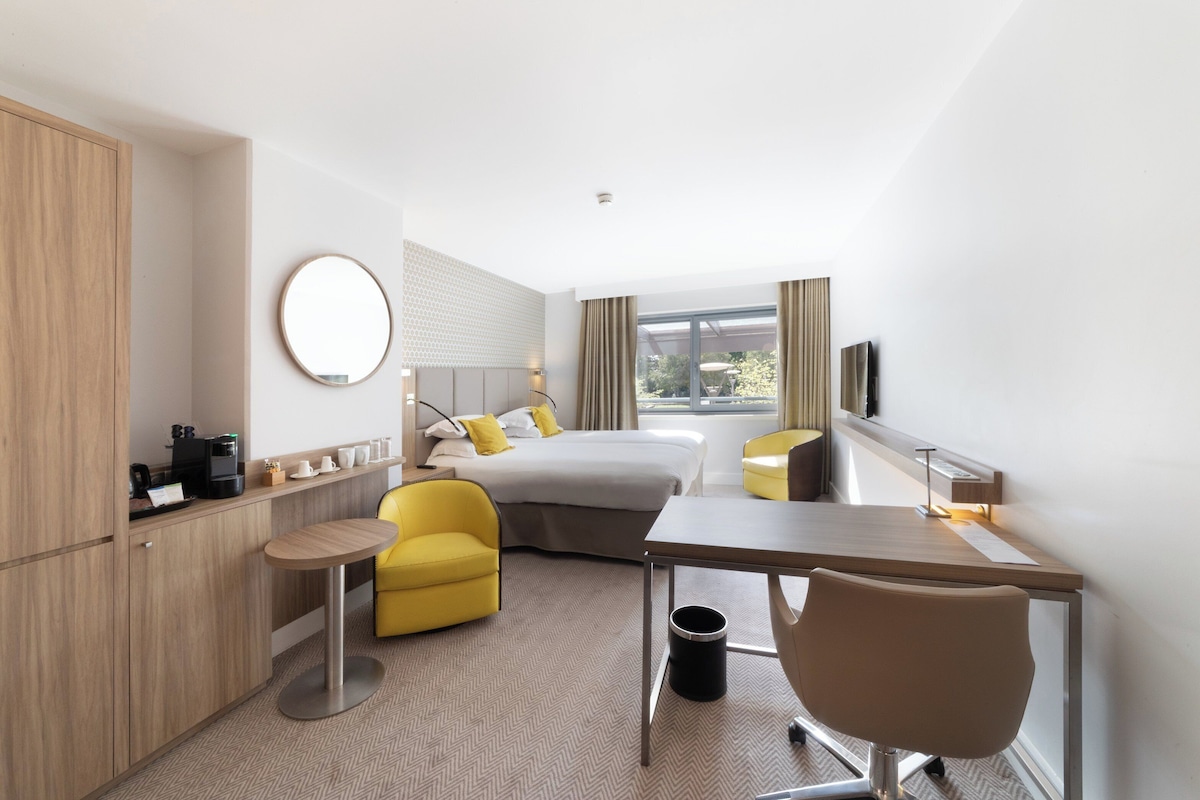 A modern hotel room with a bed, a desk, a chair, a small table, a round mirror, and a TV.