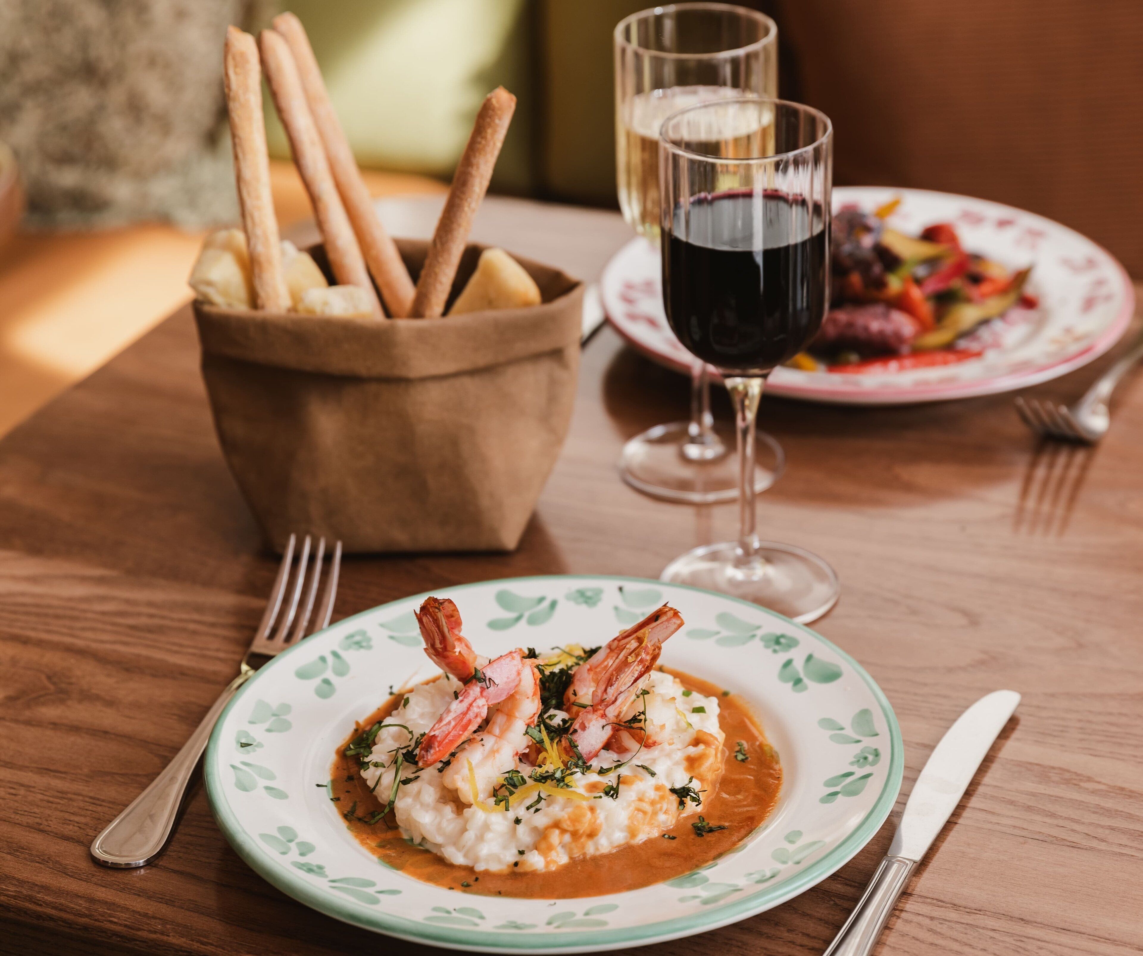 Una mesa de madera con un plato de risotto de mariscos, una copa de vino tinto y un bol de palitos de pan.