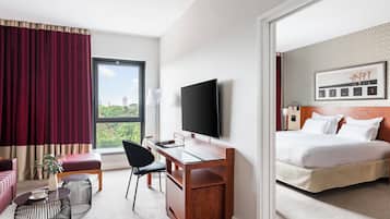 A hotel room with a bed, a desk with a TV, a chair, a small table, and a window with curtains.