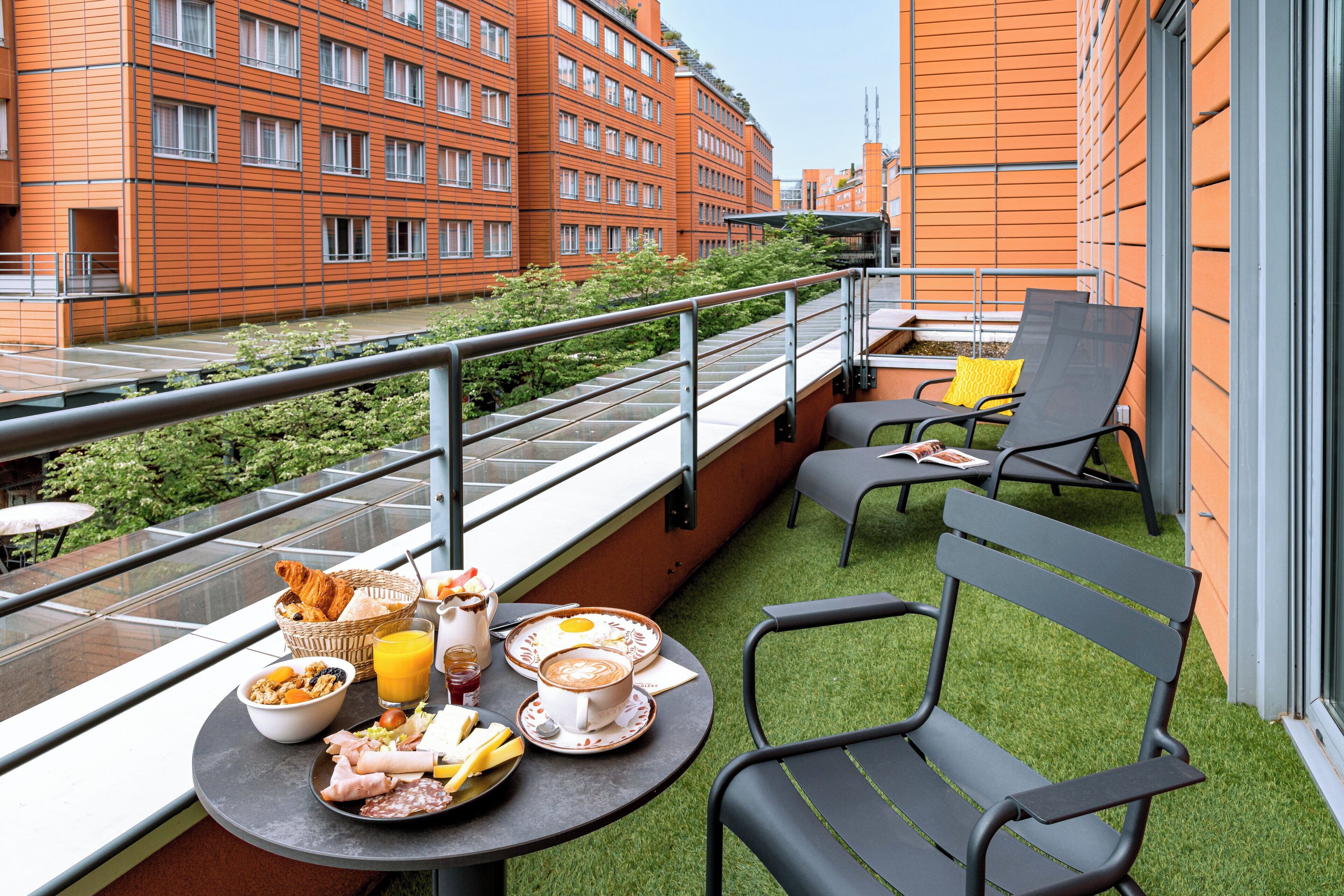 A balcony with a table set for breakfast, including croissants, a bowl of cereal, and a cup of coffee. There are two chairs and a small table with a book.