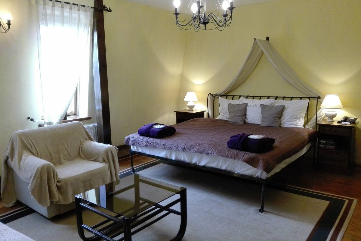 A bedroom with a bed, a chair, a glass table, and a nightstand with a lamp.