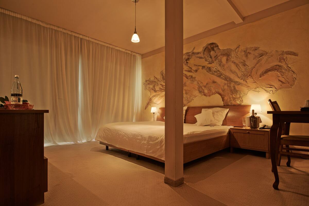 A hotel room with a large bed, a desk, a chair, a nightstand, a lamp, and a wall mural.