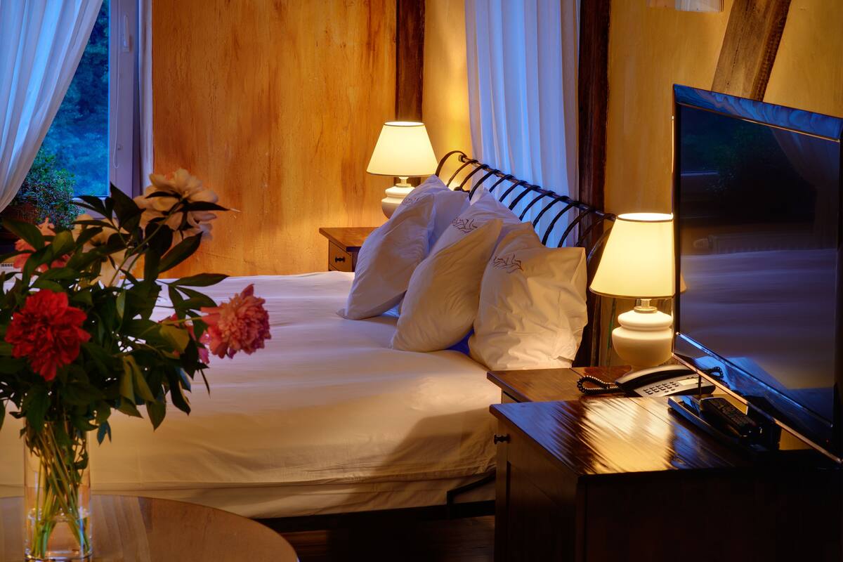 A hotel room with a bed, two bedside lamps, a TV, a telephone, and a vase with flowers.