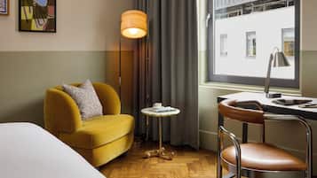 Ein Hotelzimmer mit gelbem Sessel, einem kleinen runden Tisch mit Tasse, einem Schreibtisch mit Lampe und Blick auf ein Gebäude durch ein Fenster.