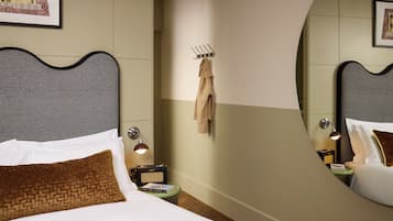 Quarto de hotel com cama, mesa de cabeceira, luminária, espelho e cabideiro fixado na parede.