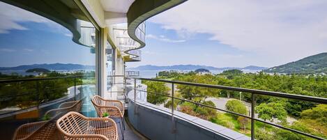Un balcón con sillas de mimbre y una mesa de vidrio con vistas a un paisaje urbano y montañas al fondo.