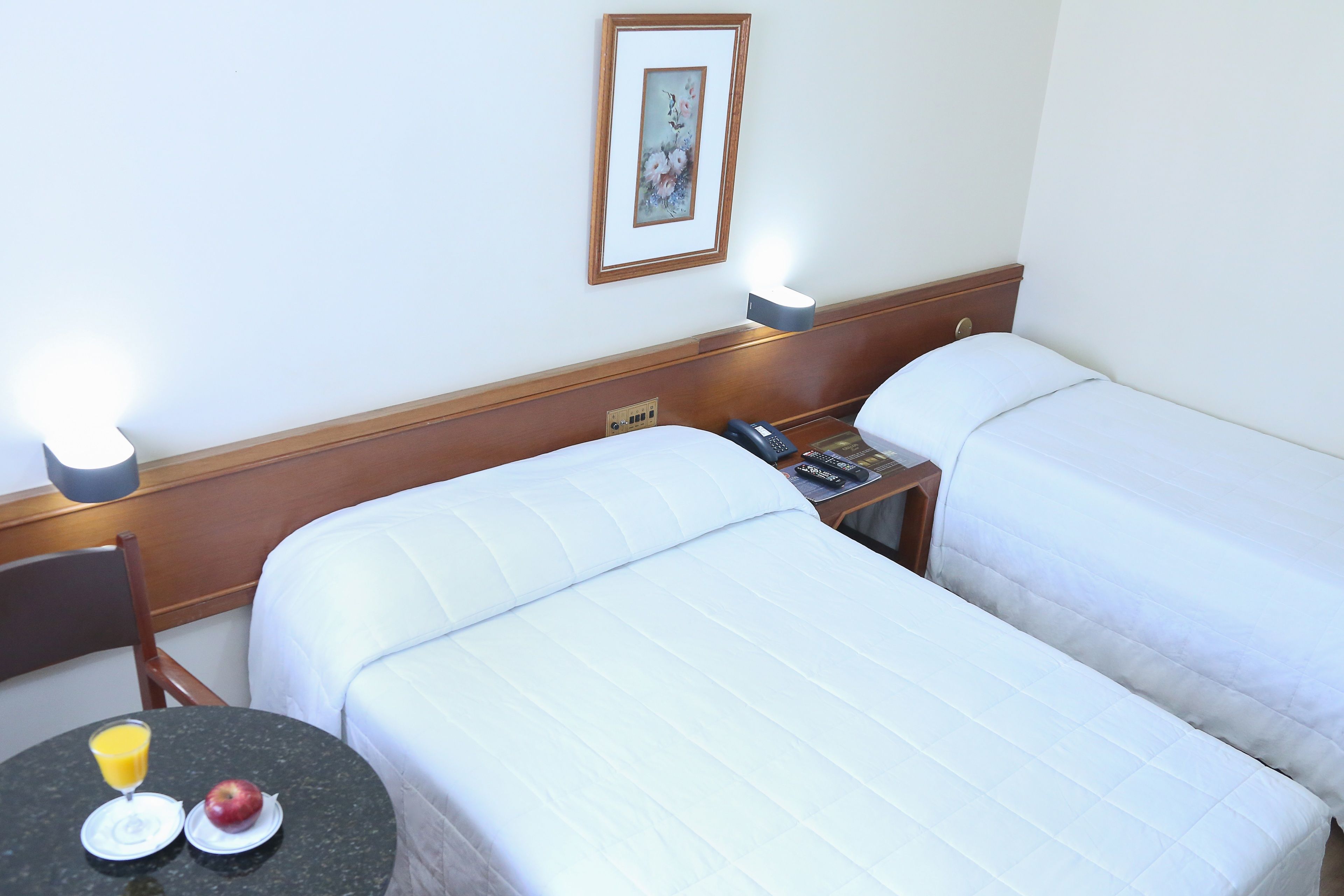 Quarto de hotel com duas camas, uma mesinha de cabeceira com telefone e abajur, e uma pequena mesa com um copo de sumo de laranja e uma maçã.