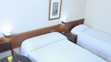 Quarto de hotel com duas camas, uma mesinha de cabeceira com telefone e abajur, e uma pequena mesa com um copo de sumo de laranja e uma maçã.