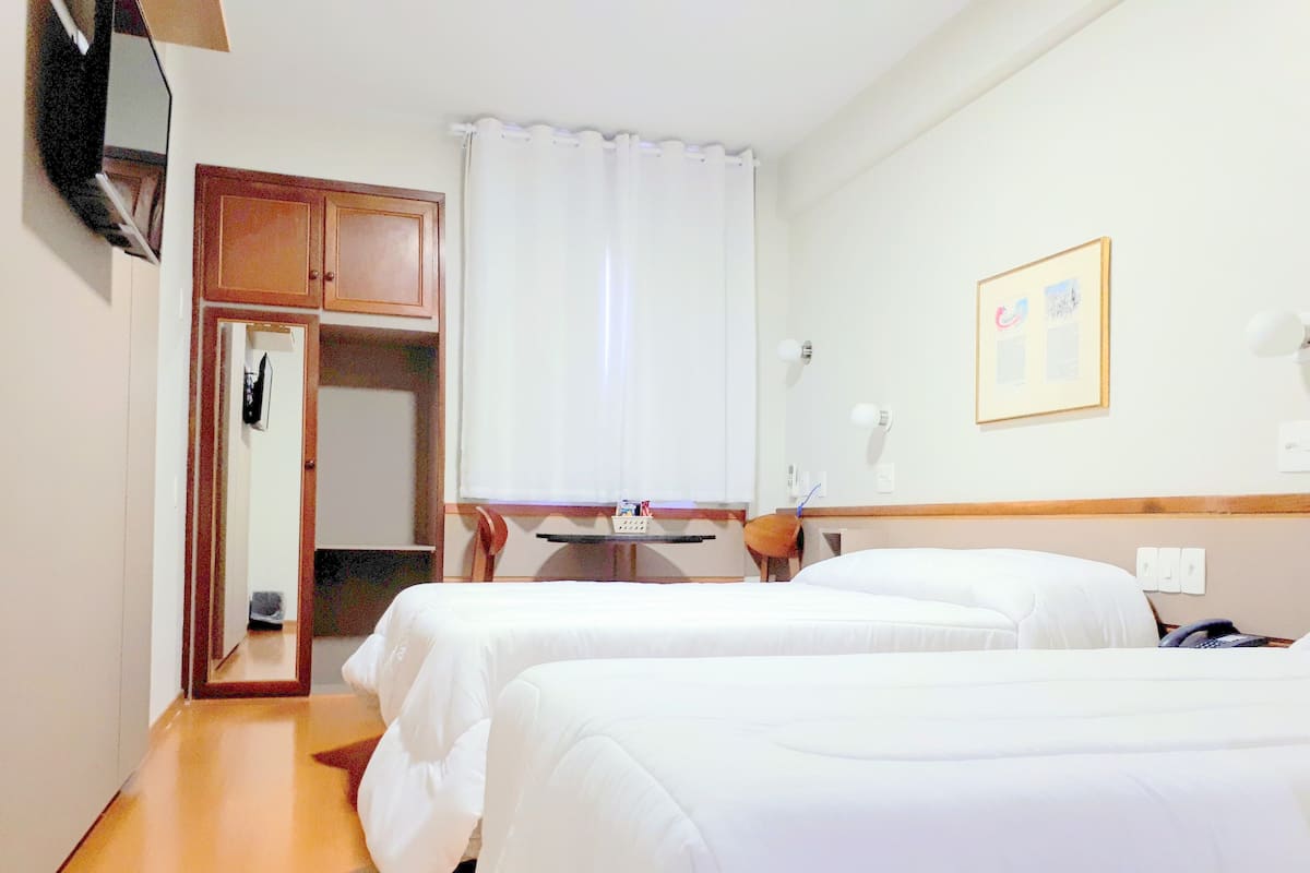 A hotel room with two single beds, a wooden wardrobe, a desk with a chair, a small table, and a mirror.