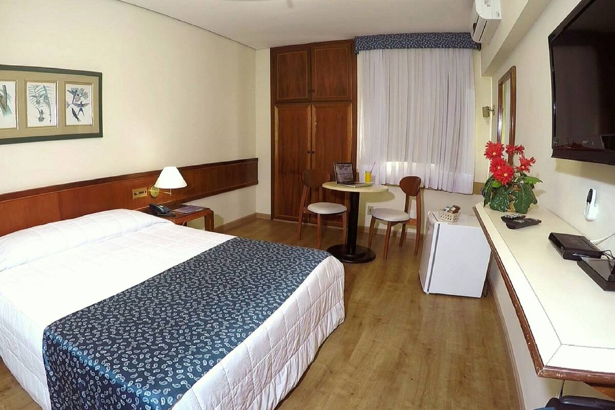 A hotel room with a bed, a desk with a computer, a chair, a small table with a lamp, and a TV.
