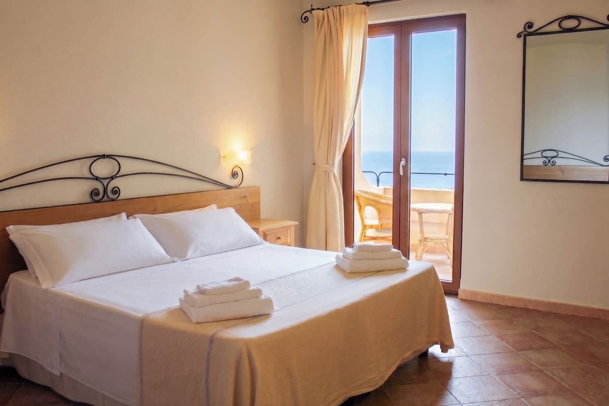 A neatly made bed with white linens, a wooden headboard, a nightstand with a lamp, and a view of the sea through a glass door.