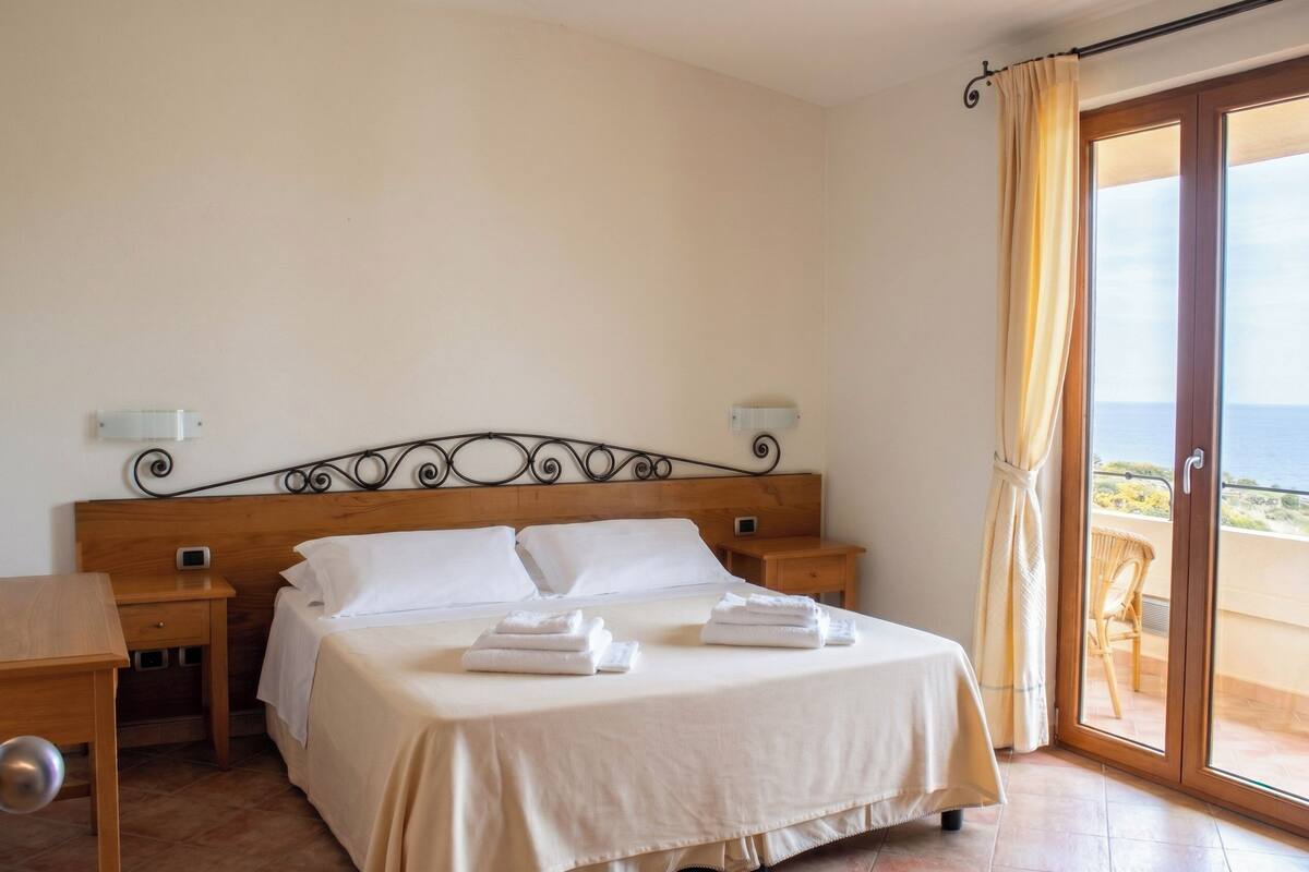 A hotel room with a bed, bedside tables, a desk, and a view of the sea through a glass door.