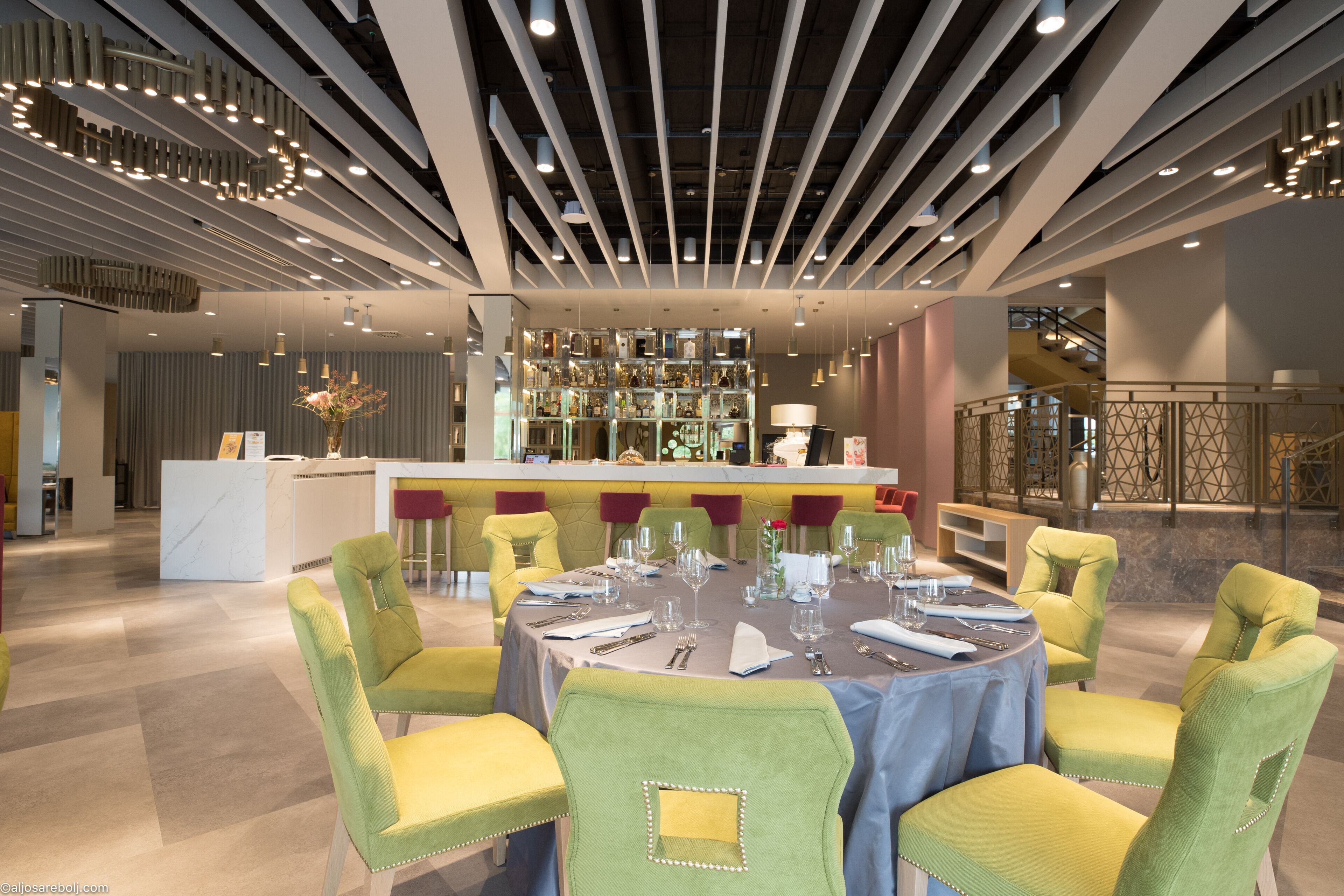 A dining area with round tables, green upholstered chairs, and a bar area in the background.
