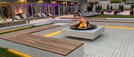 Un espacio al aire libre con sillones de descanso, un hogar a leña y un edificio moderno al fondo.