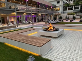 Un espacio al aire libre con sillones de descanso, un hogar a leña y un edificio moderno al fondo.