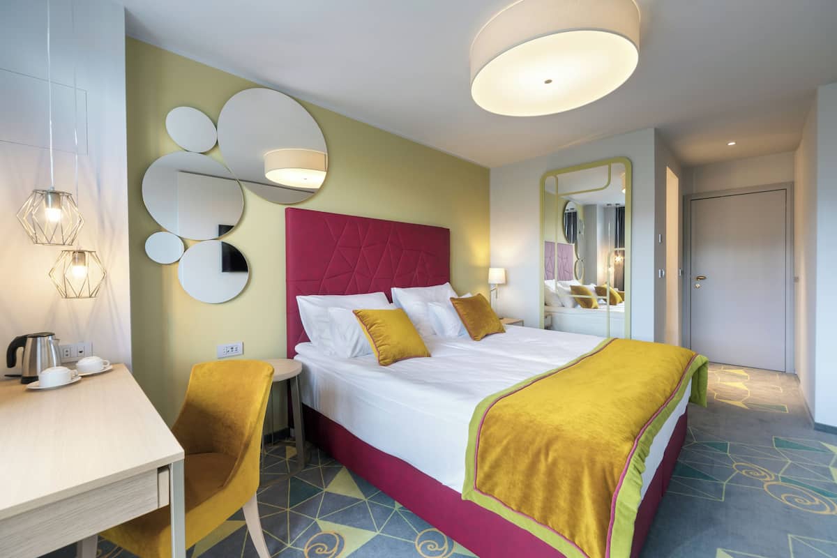 A hotel room with a large bed, a desk with a chair, a mirror, and a coffee maker.