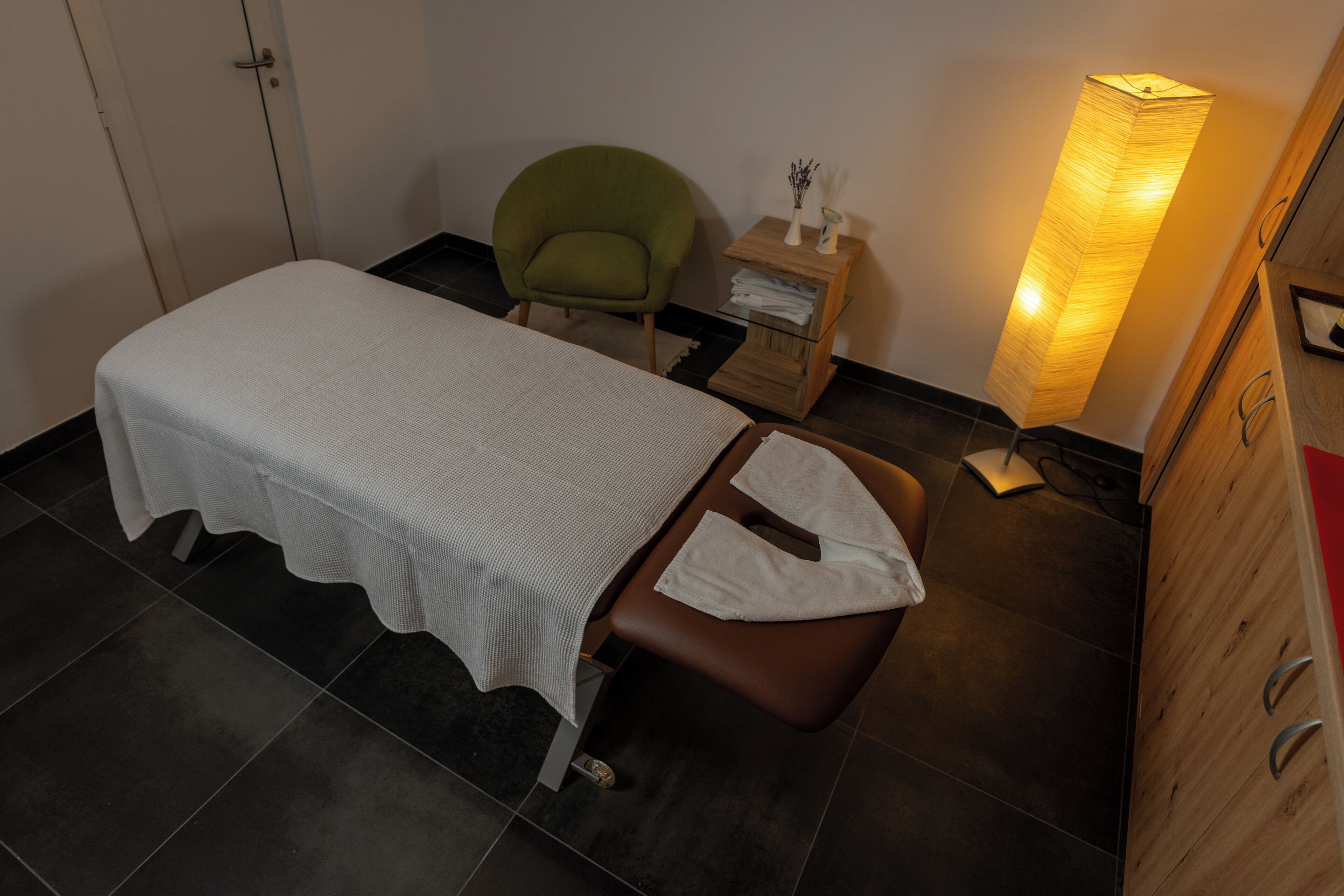 Una stanza piccola e ordinata con un letto singolo, una sedia verde, un comodino con una lampada e un armadio in legno.