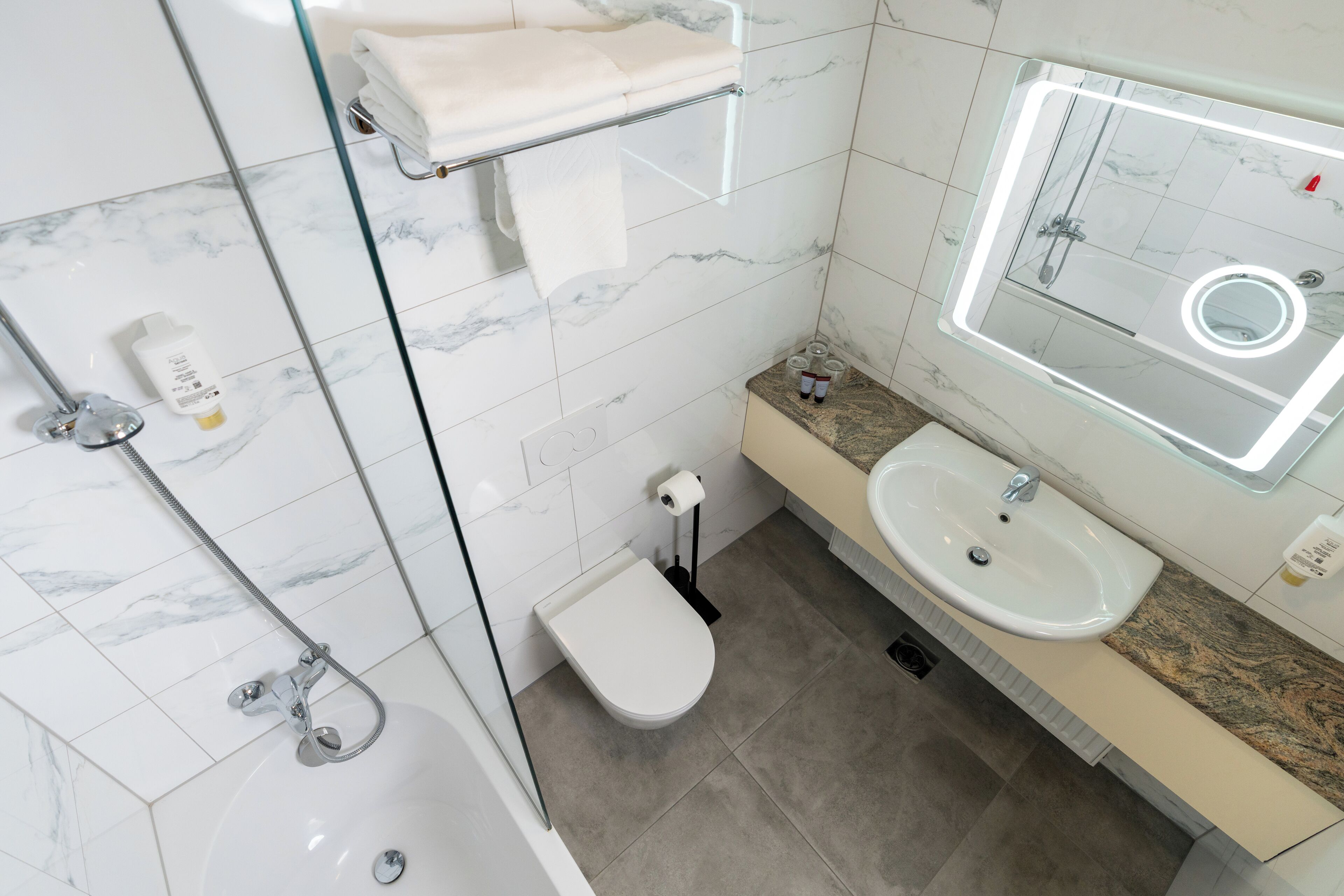 A modern bathroom with a glass-enclosed shower, a white toilet, a sink with a marble countertop, and a mirror with a built-in light.