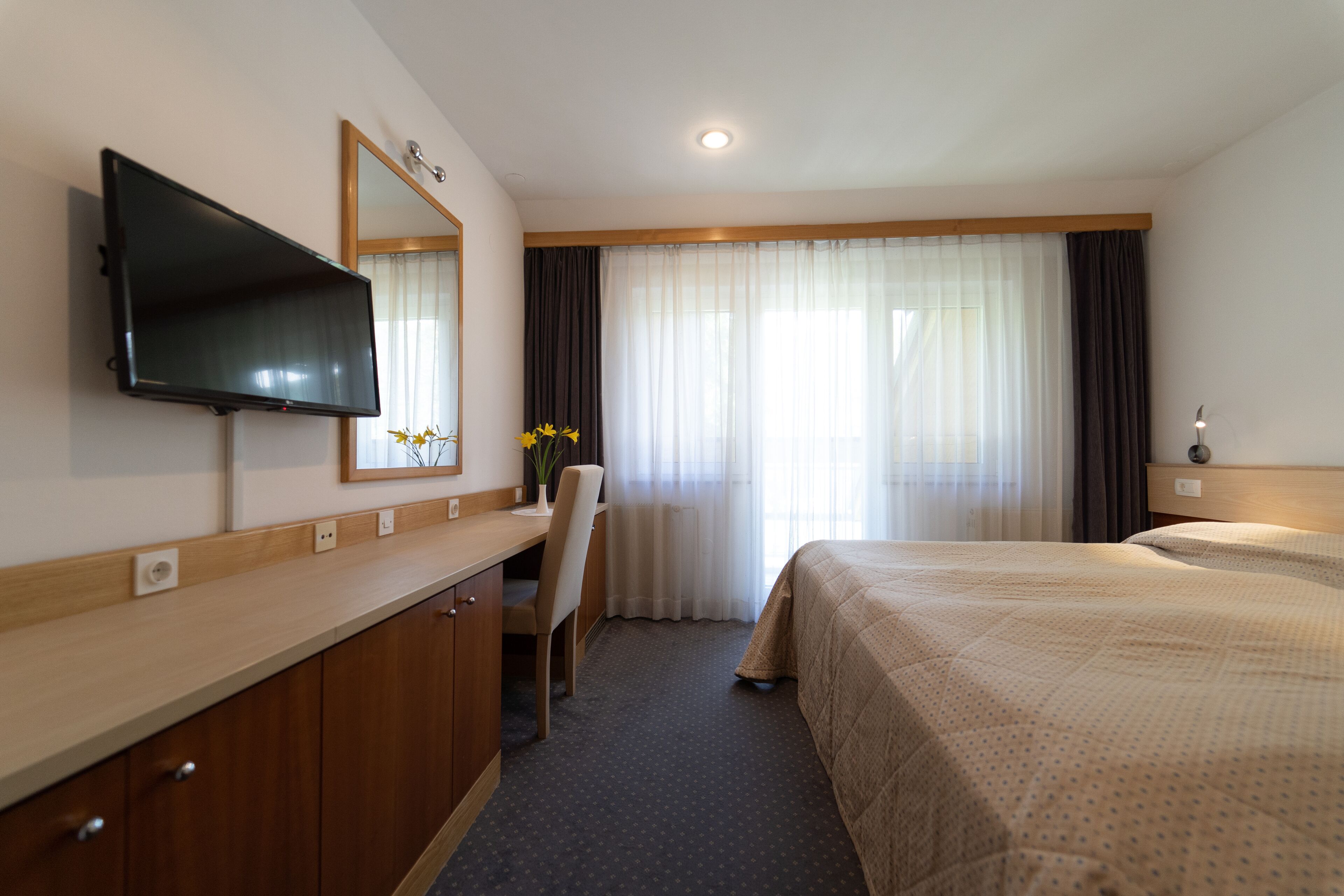A hotel room with a bed, a desk with a chair, a television, and a large window with curtains.