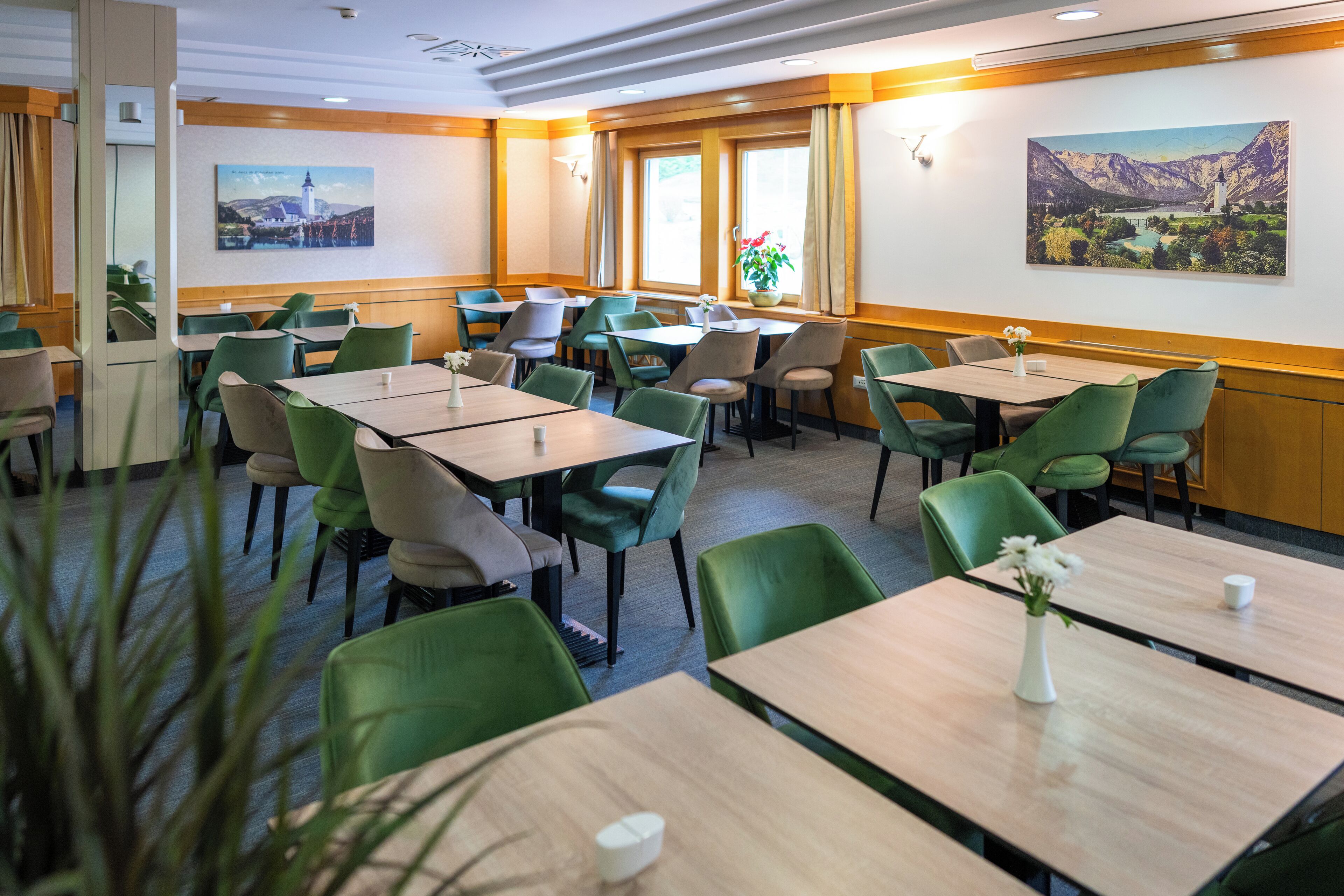A hotel dining area with multiple tables and chairs, large windows with curtains, and wall-mounted artwork.