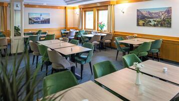 A hotel dining area with multiple tables and chairs, large windows with curtains, and wall-mounted artwork.