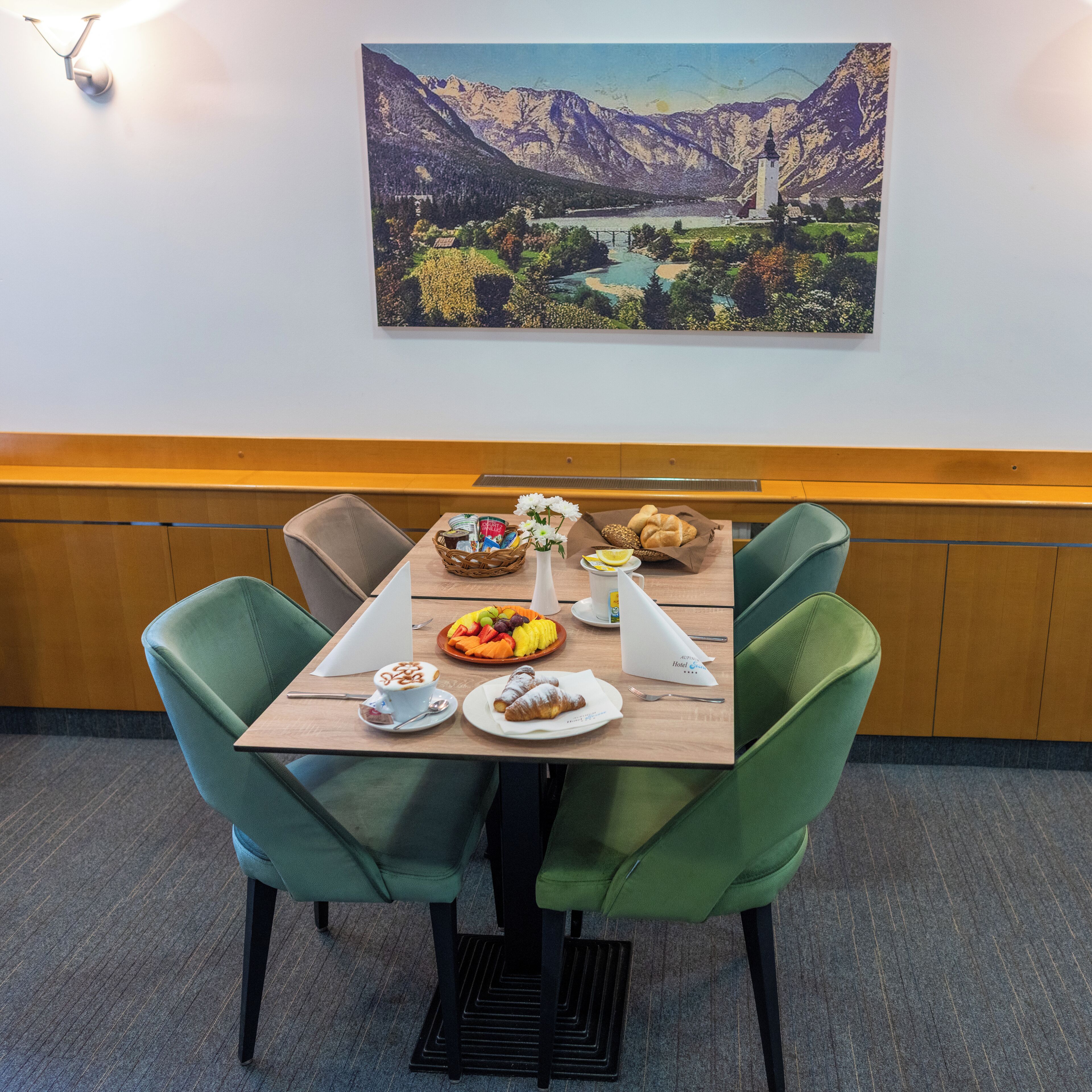 A dining table with a meal, green chairs, a painting of a mountain landscape, and a wooden bench along the wall.
