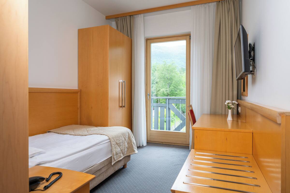 Ein Hotelzimmer mit Bett, Schreibtisch, Kleiderschrank, Fernseher und einem Fenster mit Blick auf Grünflächen.
