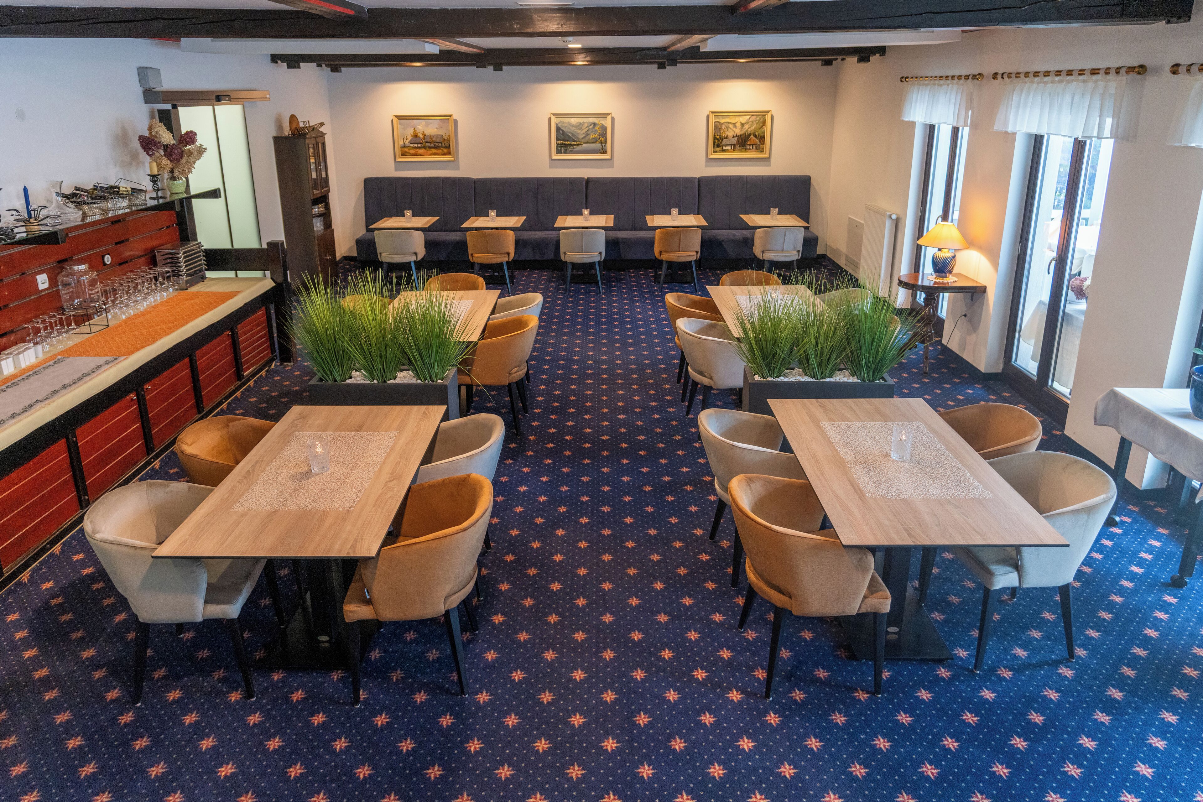 A restaurant with a blue patterned carpet, wooden tables, and chairs. There are potted plants and framed pictures on the walls.