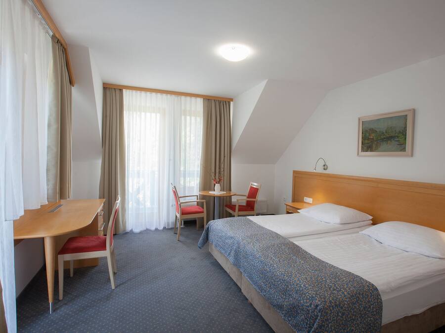 Quarto de hotel com uma cama, uma escrivaninha, duas cadeiras, uma mesinha e um quadro na parede.