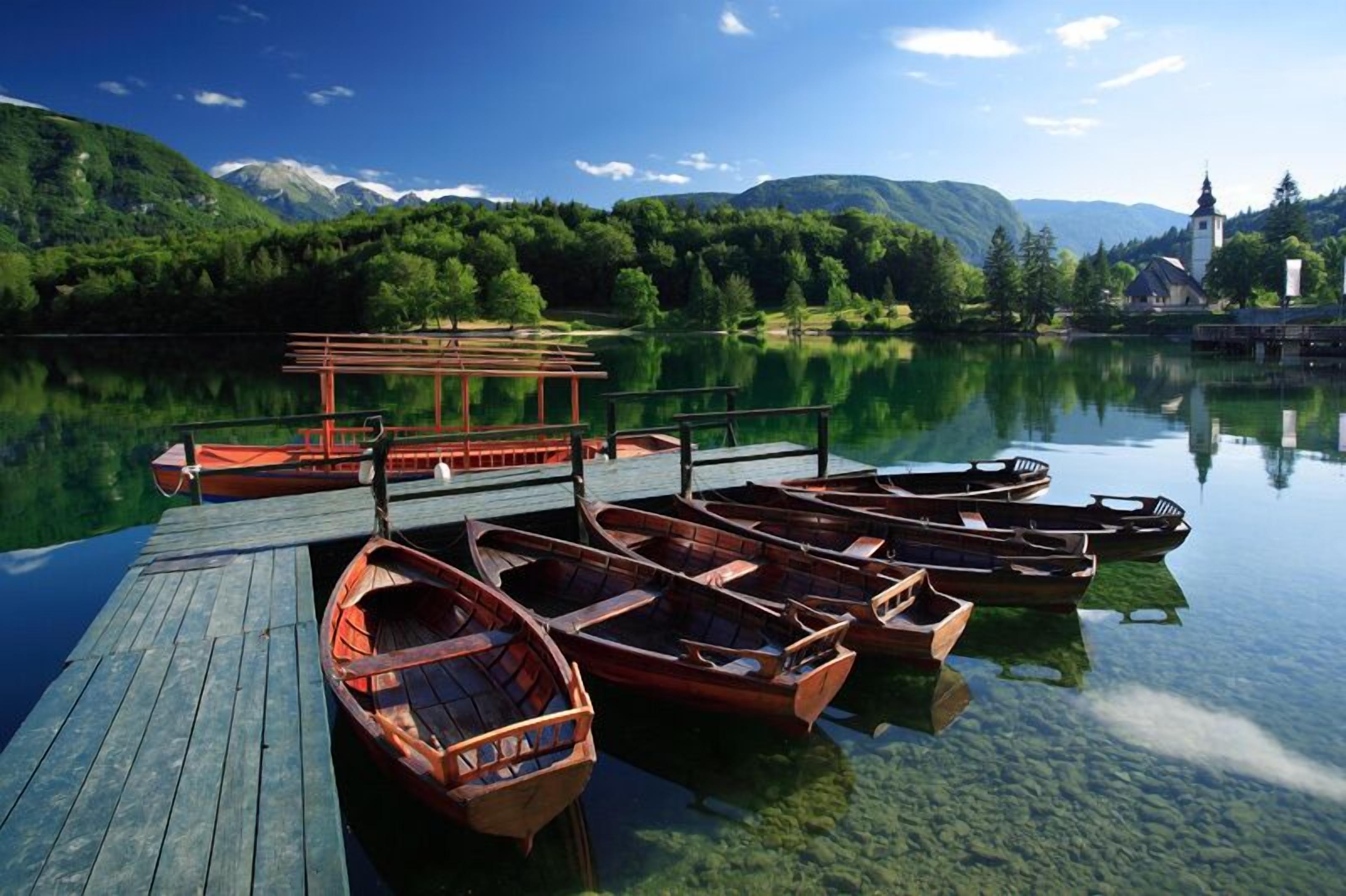 Un muelle de madera con varias lanchas y un embarcadero que se adentra en un lago tranquilo rodeado de abundante vegetación y montañas.