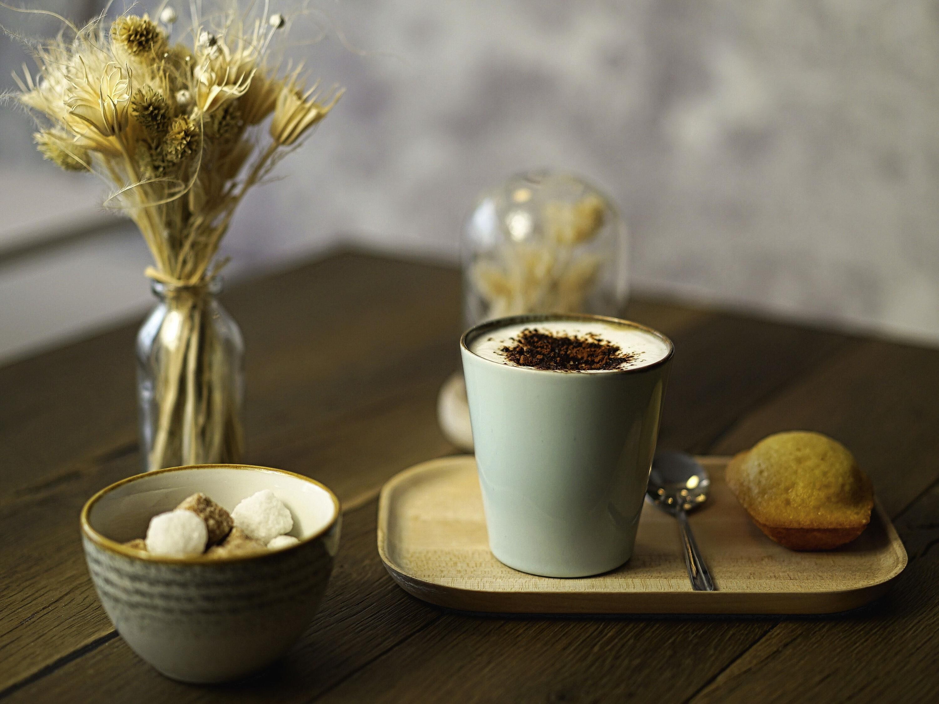 A table with a cup of coffee, a bowl of marshmallows, a muffin, and a vase with dried flowers.