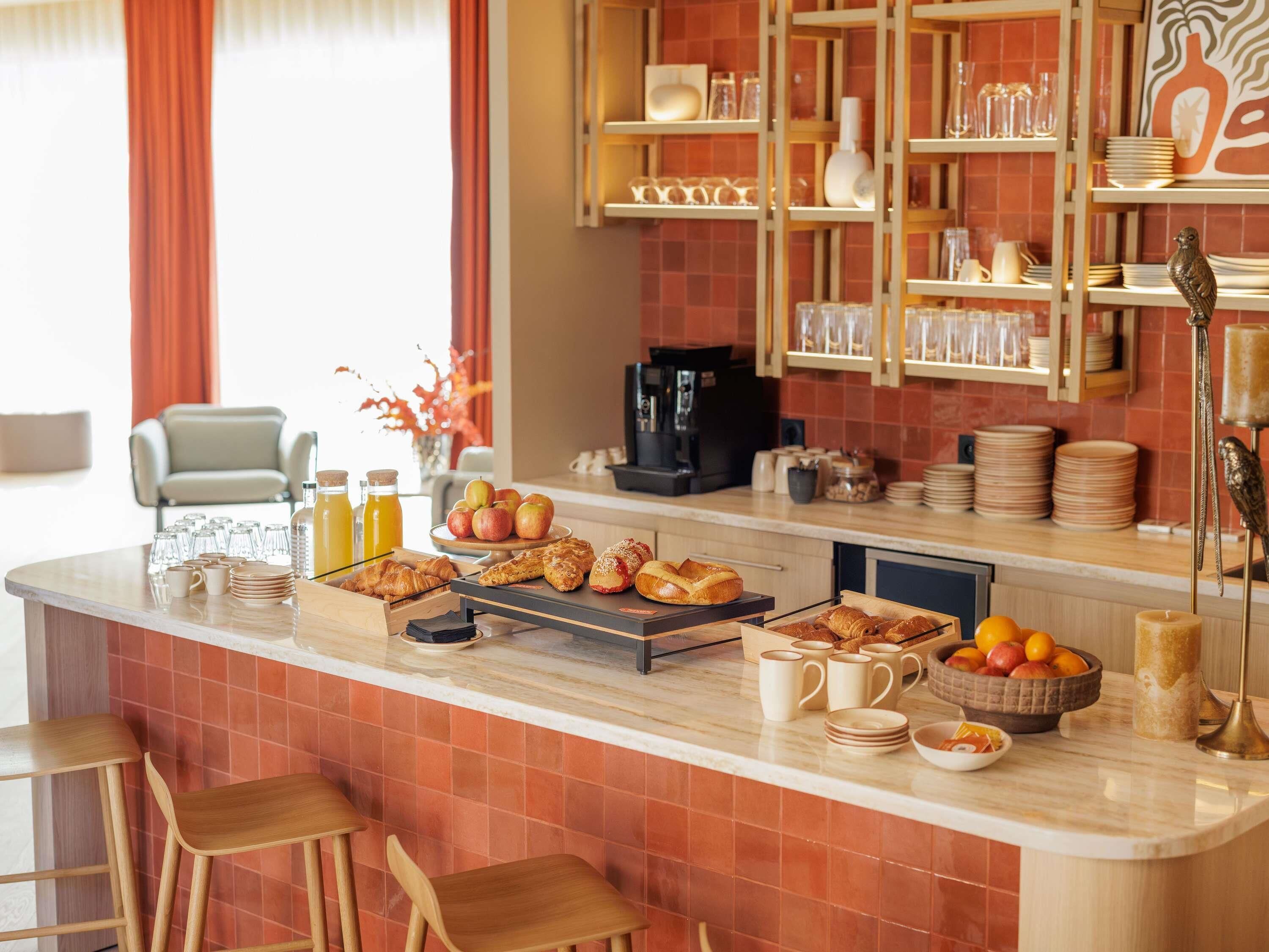 Un comptoir de petit-déjeuner avec des viennoiseries, des fruits et du café, entouré de tabourets de bar et d’une machine à café.