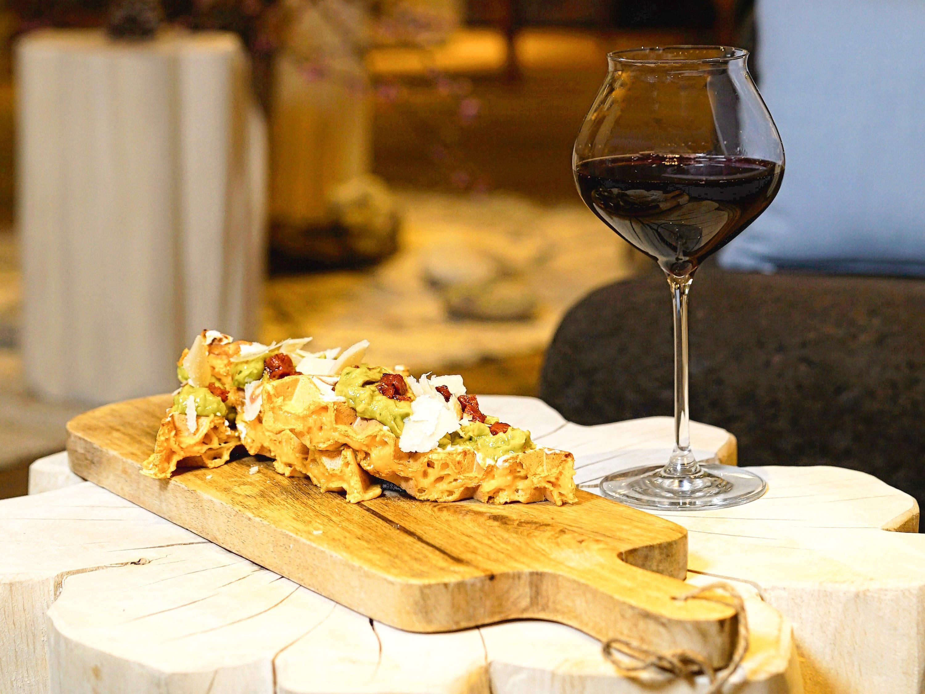A wooden serving board with a dish and a glass of red wine on a white table.