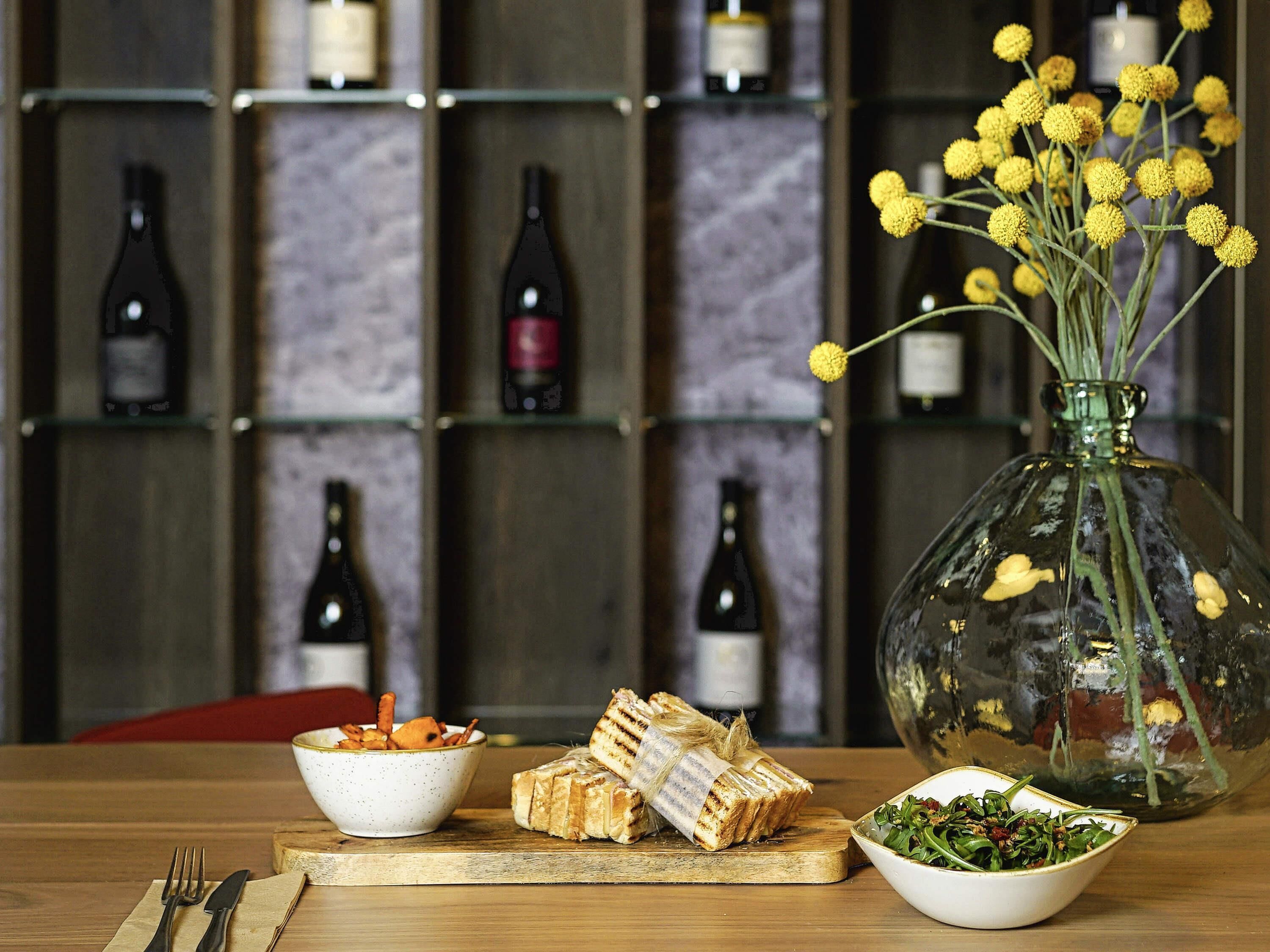 A dining table with a glass vase of yellow flowers, a bowl of greens, and a plate of sandwiches.