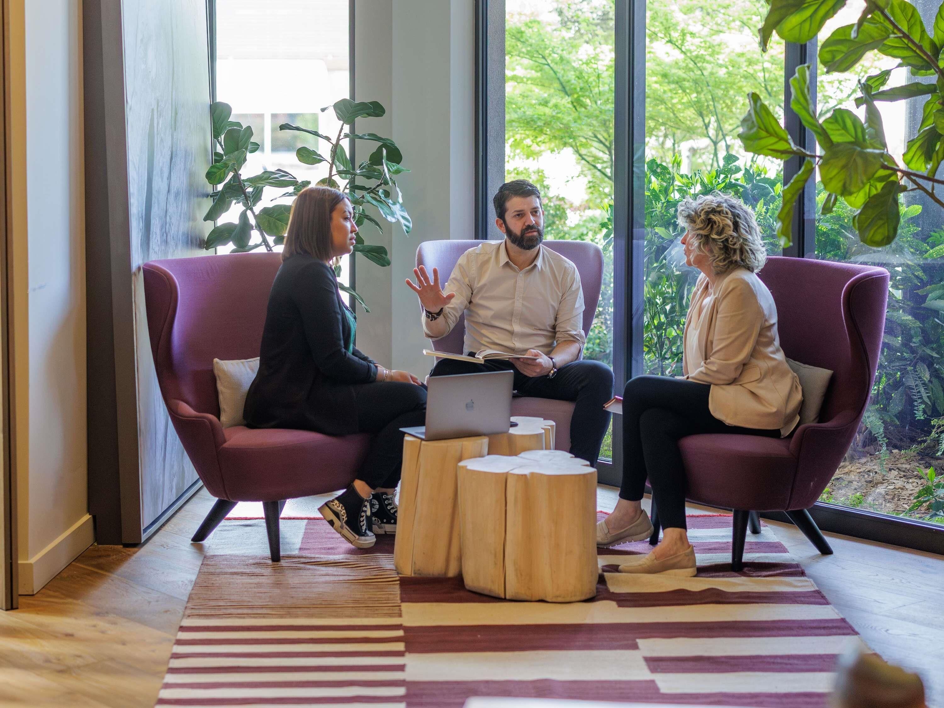 Three people seated on modern armchairs in a room with large windows, a laptop on a small wooden table, and a striped rug.