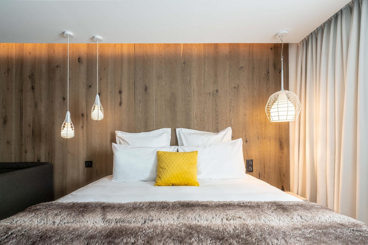 Une chambre moderne avec un mur en bois, un lit agrémenté d’oreillers blancs et jaunes, et des lampes suspendues.