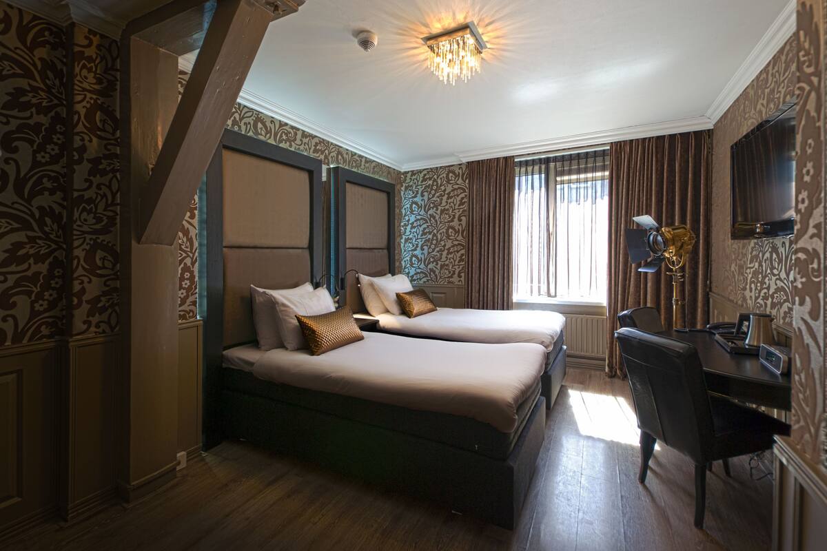 A hotel room with two beds, a desk, a chair, a TV, and a chandelier.