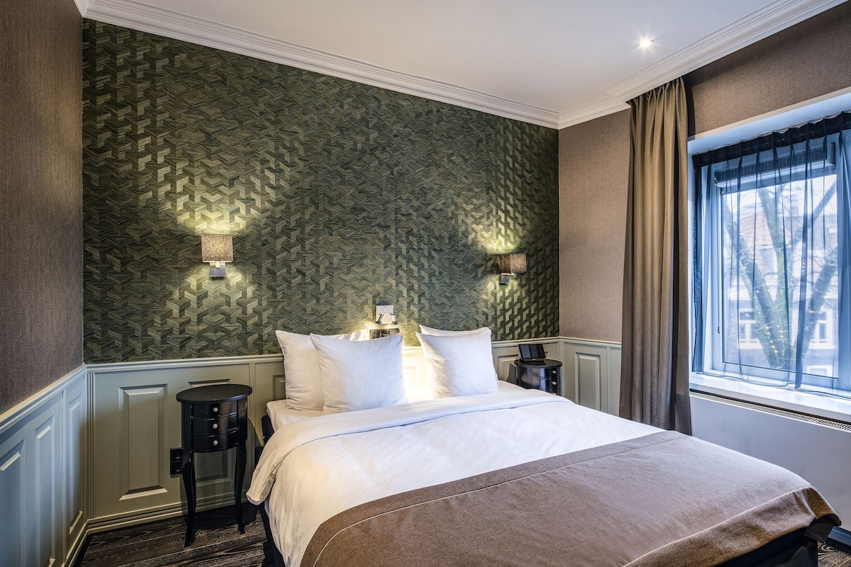 A neatly made bed with white linens and a brown throw, a nightstand with a lamp, and a patterned wallpaper on the wall.