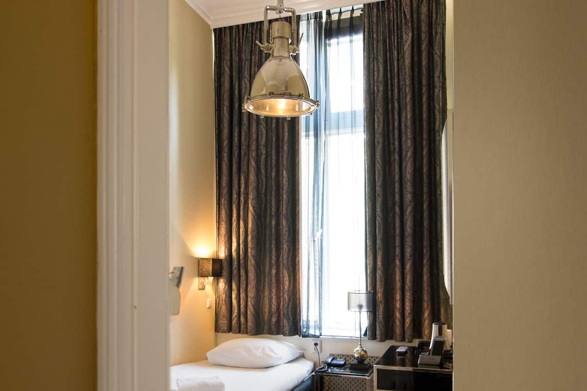 A hotel room with a bed, bedside table, lamp, and a view of the outdoors through curtains.